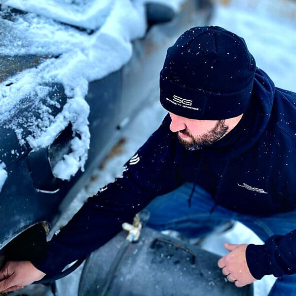 Man wearing Sargent Garage Beanie Black filling up a car tire with air in the snow.