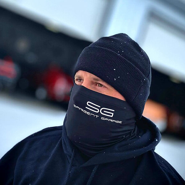 Man wearing Sargent Garage Gaiter Black in the winter with a beanie on and cars in a garage behind him.